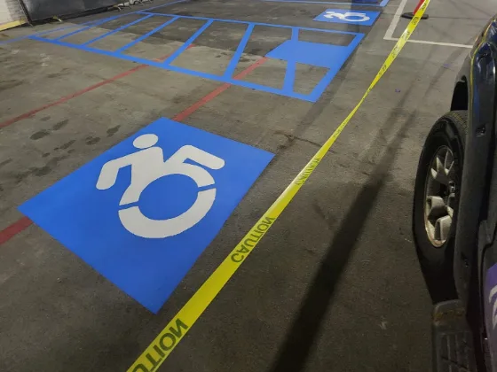 Freshly painted blue accessible-stall stencil on a parking garage deck, the hatched access aisle and blue layout lines running back between the bays with caution tape still down across the wet paint