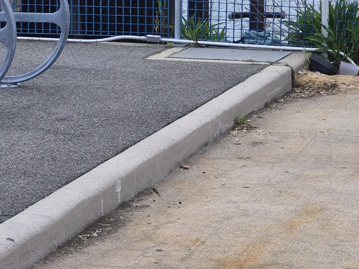 Dark asphalt parking surface meeting a concrete curb and sidewalk beside a steel fence