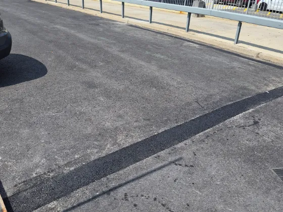 Fresh black asphalt seam repair running across a weathered commercial parking lot beside a guard rail