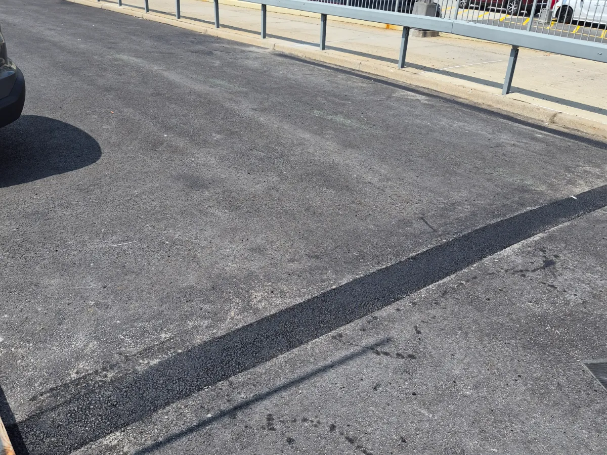 Fresh black asphalt seam repair running across a weathered commercial parking lot beside a guard rail