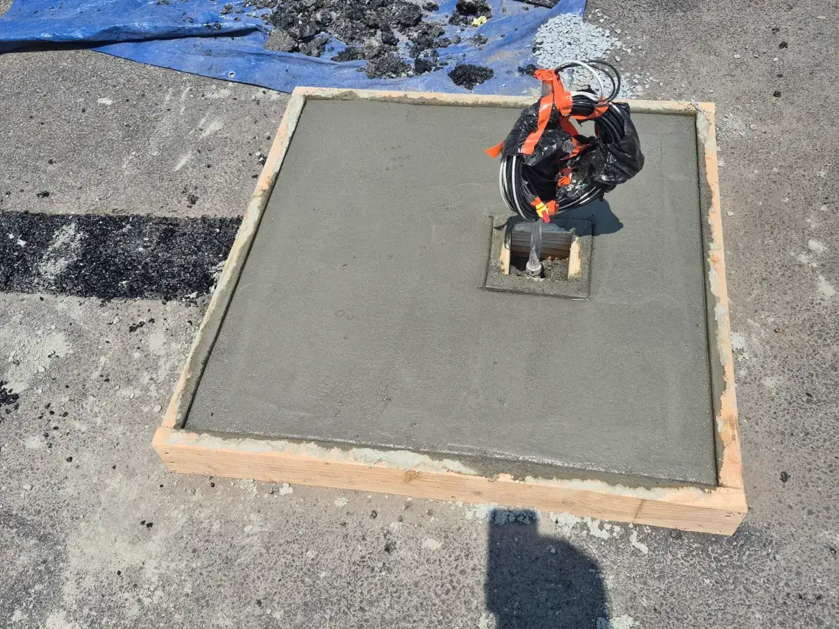 Freshly poured concrete pad inside a timber form on a parking lot, with a coiled cable conduit rising through a boxed opening
