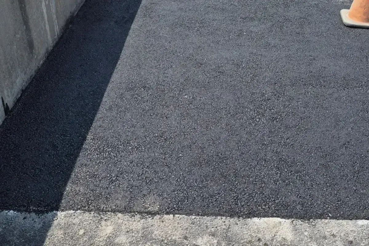 Freshly laid black asphalt patch running along a concrete wall, an orange traffic cone at its edge