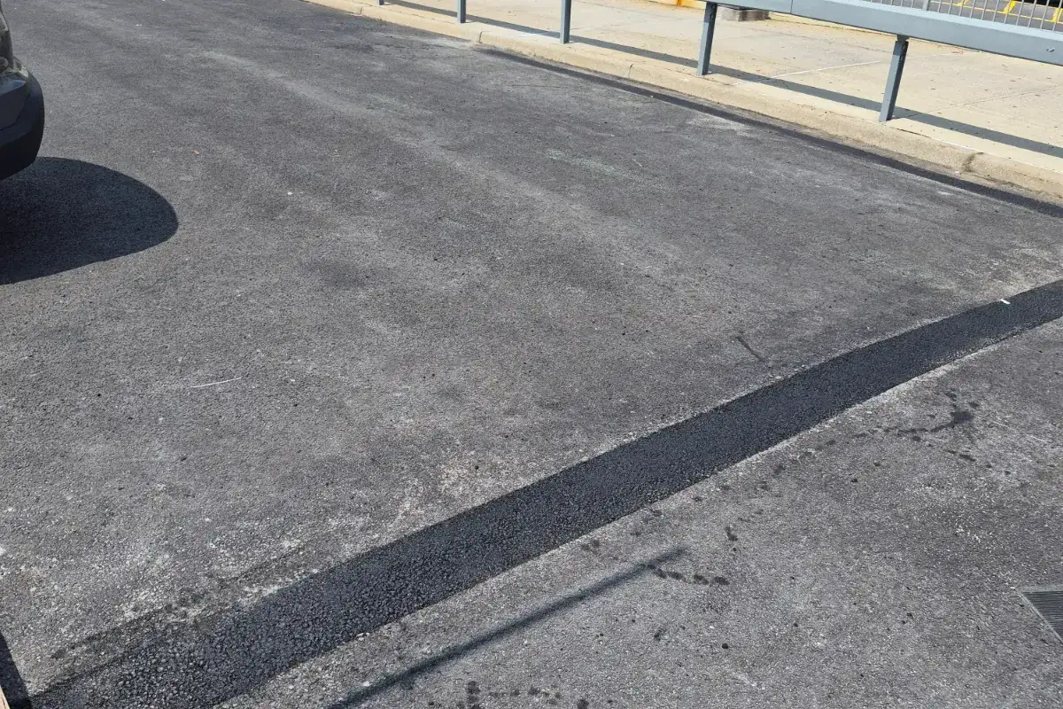 Fresh black asphalt seam repair running across a weathered commercial parking lot beside a guard rail