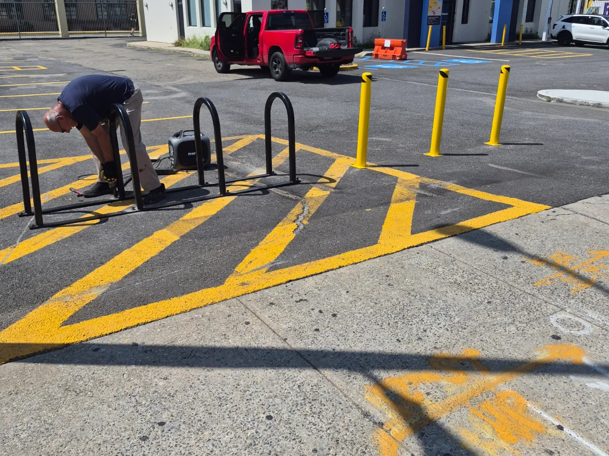 Crew member bolting down a bike rack on a commercial lot marked with fresh yellow hatching, safety bollards and accessible bays behind
