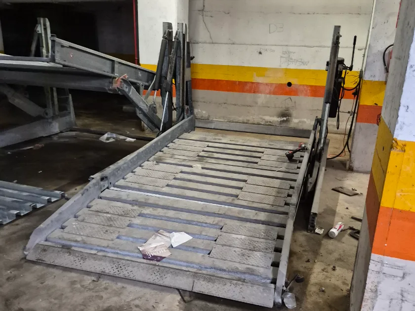 A parking stacker platform lowered in its numbered bay with the lifting posts and hydraulic ram still standing, an angle grinder resting on the deck beside it