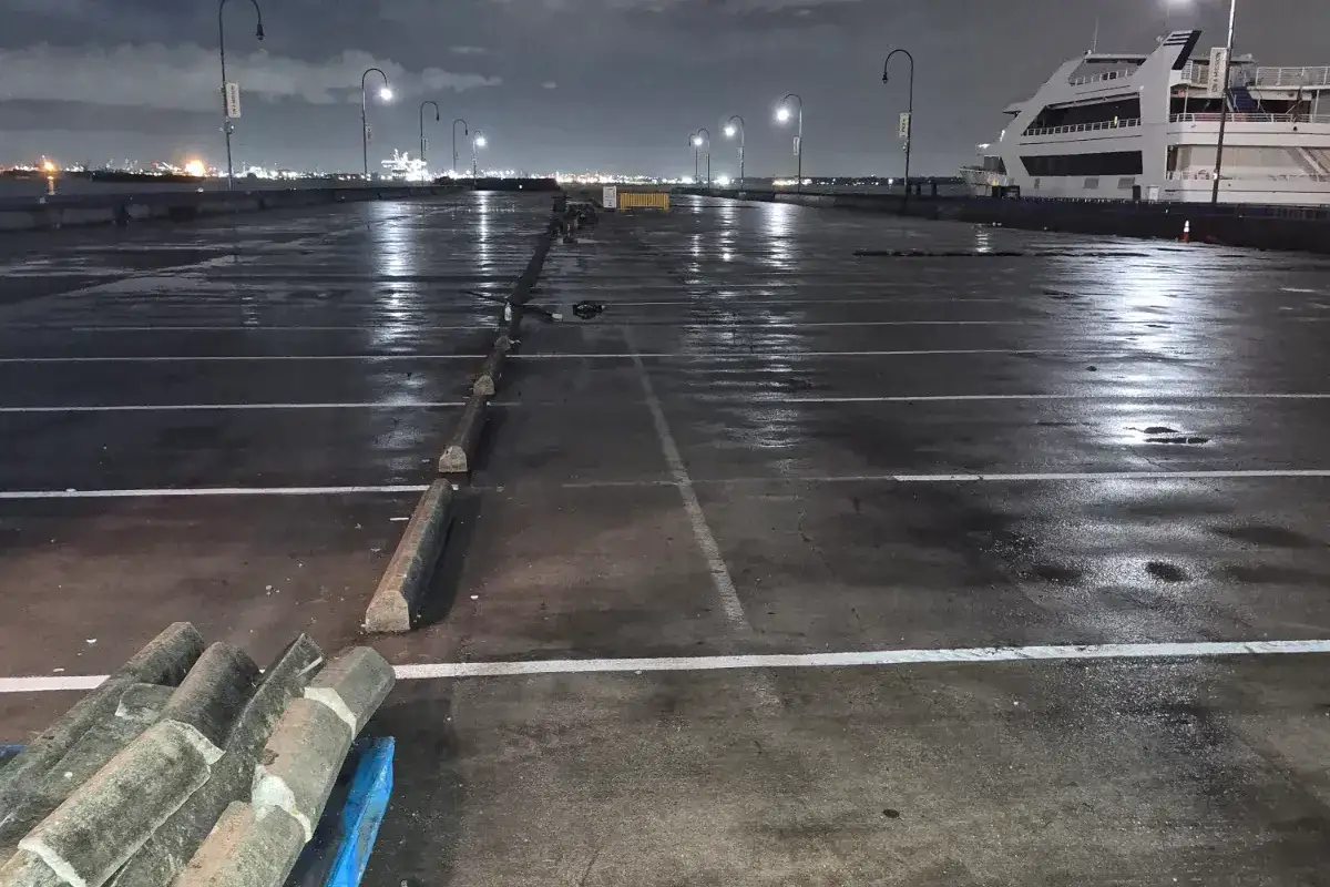 Wet waterfront parking deck at night with white stall lines, a row of wheel stops and a stack of spares in the foreground